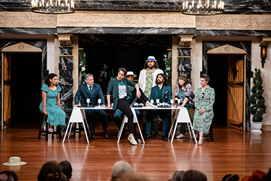 Katherine sits on a banquet table, one foot on the table top, the other hanging down to the floor. The rest of the guests sit on chairs from one end to the other behind the table. 