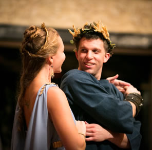 Thaisa in backless blue dress with wheat trimmings, stands behind Pericles, who is turning to look at her as he holds both her hands with his crossed arms, he in plain blue shirt and wearing a wheat garland on his head