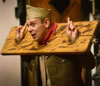 Parolles, in brown army uniform with sky cap, head andhands in stocks while standing up