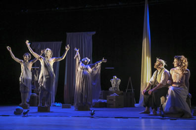 Prospero, whearing his crown, white shirt, brown vest, and ankle-high brown pants sits tot he side with Miranda, flowing hair, gold corset and white dress as they watch the gods who appear as three statues, hands uplifted, standing on pedestals. Int he background are three white sheets hanging from rods and a muscular torso maniquen.