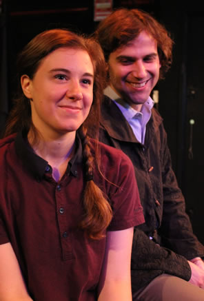 The messenger in short-sleeve brown button-down shirt and pig tales sits looking forward as Henry sits behind her smiling gently, wearing a simple winter jacket and blue oxford shirt