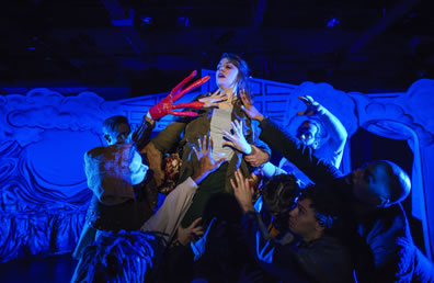 Imogen in gray blouse and bran jacket is lifted by the ensemble as others reach up to her, including Iachimo with long reg fingers