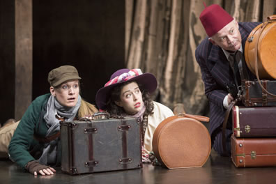 Rosalind in green jacket, blue scarfe, and coruroy breen billed gap, Celia in white dress and purple floppy hate with a circle of roses on the crown, and touchestown in red Shriners hat and pinstriped suit hide behind luggage, the curtains of Arden in the background.